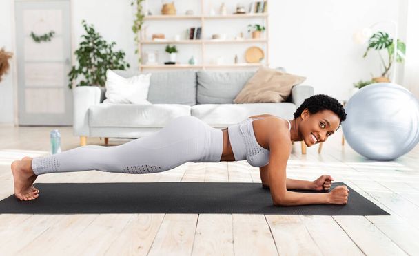 Όμορφη μαύρη γυναίκα κάνει προπόνηση γυμναστικής σε αθλητικό χαλάκι στο σπίτι, στέκεται στη σανίδα αγκώνα, ενίσχυση των μυών ABS - Φωτογραφία, εικόνα