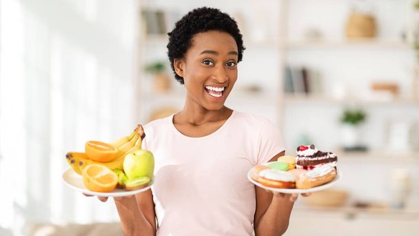 Ευτυχισμένη μαύρη κυρία κρατώντας πιάτα με φρούτα και επιδόρπια στο σπίτι, κάνοντας επιλογή μεταξύ υγιεινών και ανθυγιεινών τροφίμων - Φωτογραφία, εικόνα