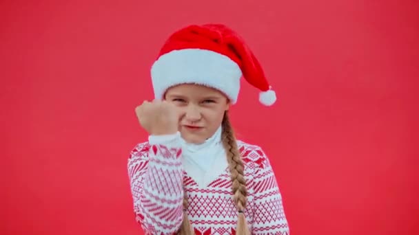 dühös lány télapó kalap és karácsonyi pulóver mutatja öklöt elszigetelt piros - Felvétel, videó