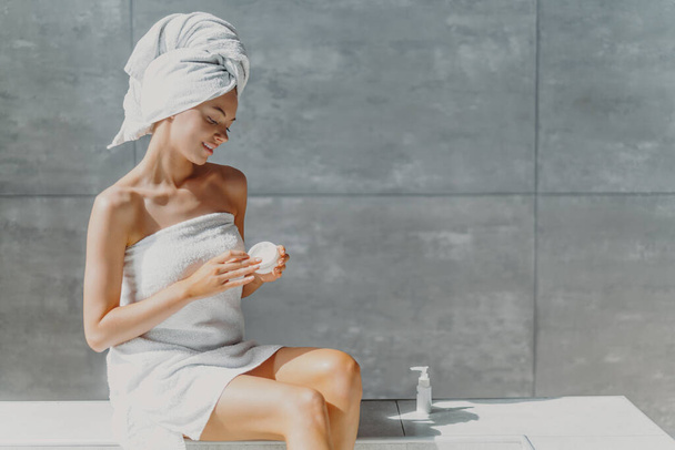 Horizontal shot of relaxed beautiful womam applies body lotion and nourishing cream after shower, poses in bathroom against grey wall, sits wrapped in towel, undergoes beauty treatments. Spa concept - Фото, зображення