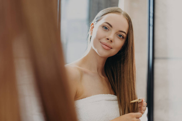 Pretty young woman with minimal makeup, full lips looks at herself in mirror, combs long straight hair, wrapped in bath towel, cares about herself, wants to have fabulous look. Hair care concept - Photo, Image