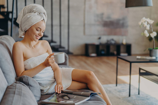 Horizontal shot of relaxed satisfied woman has healthy skin reads magazine poses with cup of coffee or tea at couch wrapped in soft towels enjoys spending time at home. Refreshment leisure spa - Photo, Image