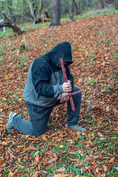 Un hombre en una capucha, inclinando la cabeza, de pie sobre una rodilla en oración y sostiene una cruz debajo. - Foto, imagen