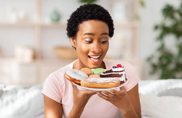 Diet breakdown and unhealthy nutrition concept. Young African American lady holding plate of sweet pastries at home - Фото, изображение
