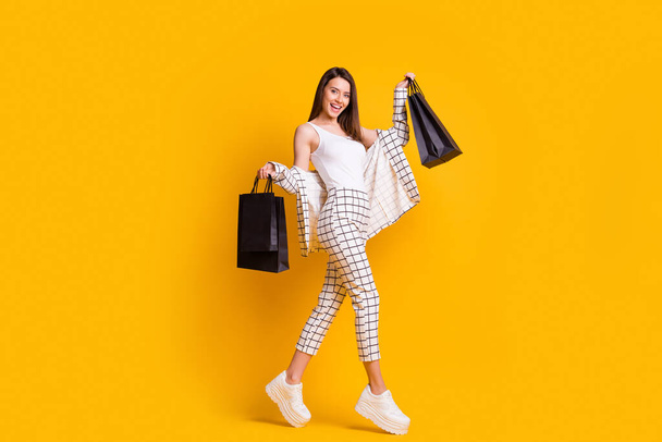 Full body profile photo of brown hair girl jump go shopping hold black bags wear white singlet plaid pants isolated over yellow color background - Zdjęcie, obraz