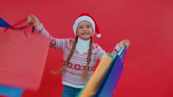Noel Baba şapkalı ve Noel süveterli kız kırmızı üzerinde izole edilmiş alışveriş torbalarıyla dans ediyor. - Video, Çekim