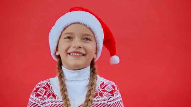 chica en sombrero de santa y suéter de Navidad jugando con trenzas aisladas en rojo - Metraje, vídeo