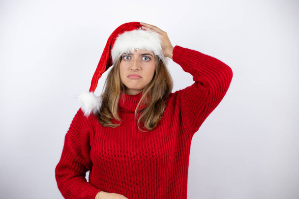 Μια νεαρή όμορφη ξανθιά γυναίκα φοράει ένα κόκκινο καθημερινό πουλόβερ και ένα χριστουγεννιάτικο καπέλο πάνω από λευκό φόντο βάζει το ένα χέρι στο κεφάλι της χαμογελώντας σαν να είχε ξεχάσει κάτι. - Φωτογραφία, εικόνα