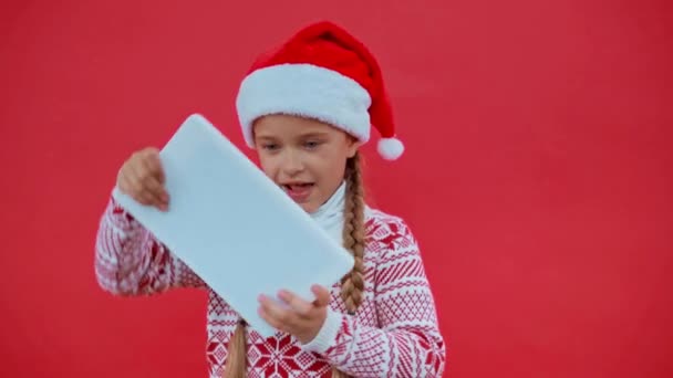 chica en sombrero de santa y suéter de Navidad jugando en la tableta digital aislado en rojo - Imágenes, Vídeo