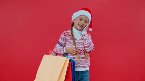 dívka v Santa klobouk s nákupními taškami mluví na smartphone izolované na červené - Záběry, video