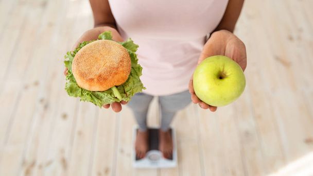 Above view of black woman holding apple and hamburger, standing on scales, making choice of her diet, closeup - 写真・画像