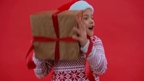 взволнованная девушка в шляпе Санты и рождественский свитер трясущий подарочную коробку изолированы на красный - Кадры, видео