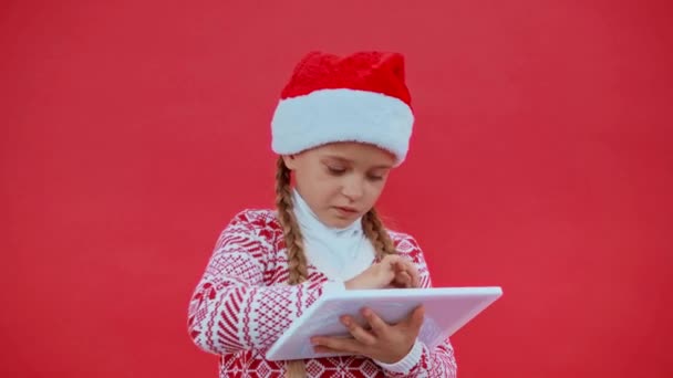 girl in santa hat and christmas sweater using digital tablet isolated on red - Footage, Video