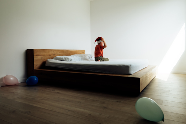 criança na cama dos pais com chapéu de Papai Noel e balões celebrando feriado de Natal em tempo de bloqueio pandêmico - Foto, Imagem
