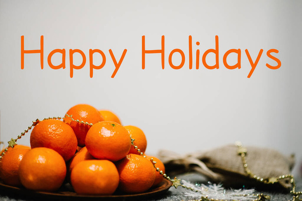 Text Happy Holidays. Tangerines in plate and rustic stylish sack on gray knitted background. space for text, design. Greeting Festive Card. Merry Christmas Happy New Year's 2019 Celebration Concept. - Fotó, kép