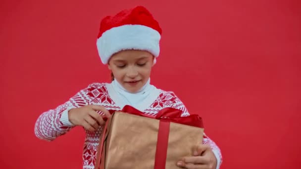 dziewczyna w Santa kapelusz i Boże Narodzenie sweter otwarcie pudełko prezent odizolowany na czerwono - Materiał filmowy, wideo