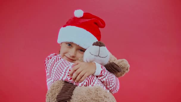 girl in santa hat and christmas sweater hugging teddy bear isolated on red - Footage, Video