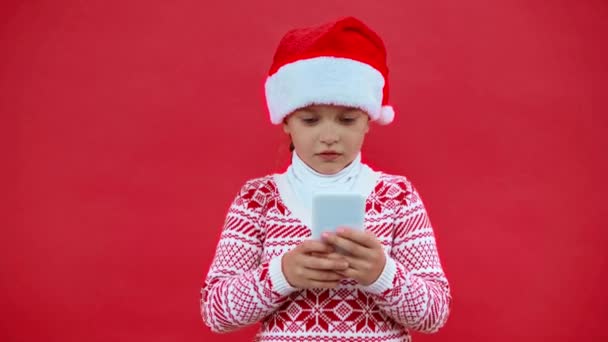 サンタ・ハットの女の子と赤で隔離されたスマートフォンを使ったクリスマスセーター - 映像、動画