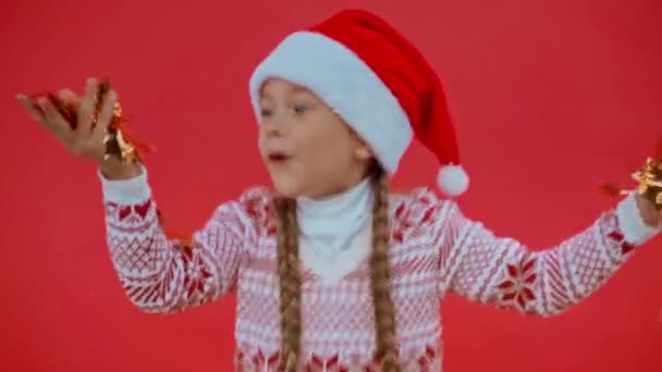Ευτυχισμένο κορίτσι με καπέλο Σάντα και χριστουγεννιάτικο πουλόβερ φυσώντας κομφετί απομονωμένο στο κόκκινο - Πλάνα, βίντεο