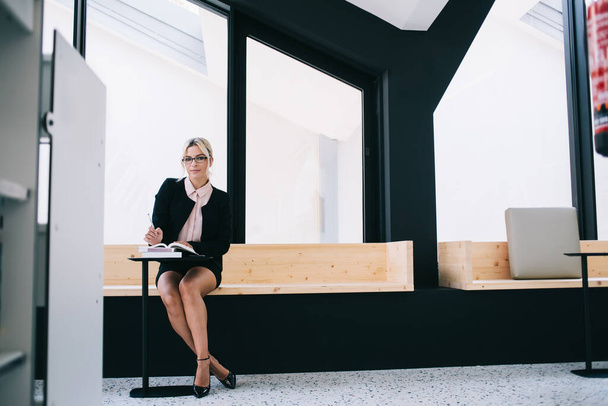 Portrait of confident female lawyer sitting in modern workshop near desk with literature books, elegant Caucasian woman in formal wear and optical glasses for eyes correction looking at camera - Foto, immagini