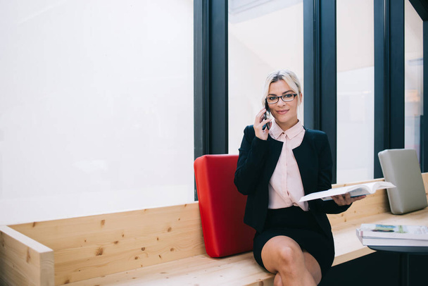 Confident smiling young female entrepreneur in black business suit holding organizer and talking on mobile phone while sitting at workspace - Photo, image