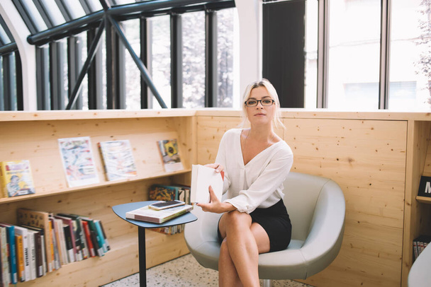 Νεαρή κομψή πολυάσχολη γυναίκα με γυαλιά ηλίου και λευκή μπλούζα με μαύρη φούστα που κοιτάζει την κάμερα ενώ κάθεται στην καρέκλα με το βιβλίο στο σύγχρονο χώρο εργασίας με ξύλινη βιβλιοθήκη - Φωτογραφία, εικόνα