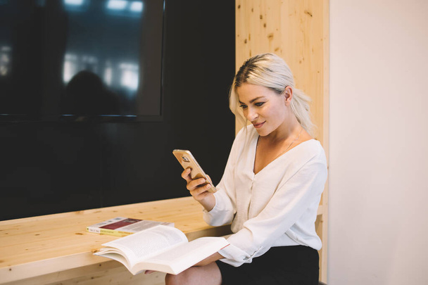 Happy young businesswoman taking photo of book pages on smartphone while sitting during work at wooden counter in modern conference room - Foto, imagen
