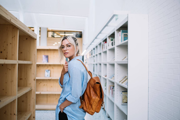Half length portrait of intelligent female reader in optical eyewear for vision protection looking at camera during time for shopping in public bookstore, beautiful woman with backpack posing indoors - Foto, afbeelding