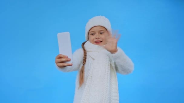 Tyttö neulottu valkoinen talvi asu heiluttaa käsi videopuhelun aikana eristetty sininen - Materiaali, video