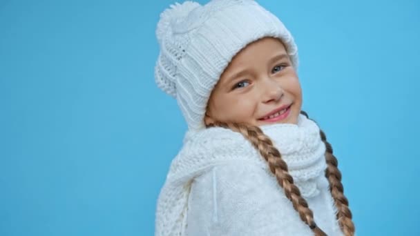 χαμογελαστό χαριτωμένο κορίτσι σε πλεκτό λευκό χειμωνιάτικο ντύσιμο απομονωμένο σε μπλε - Πλάνα, βίντεο