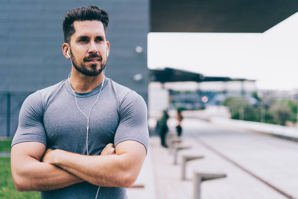 Handsome male runner dressed in active wear and electronic earphones pondering on healthy workout,Caucasian muscular trainer with crossed hands using headphones for listening motivation music playlist - Foto, Imagen