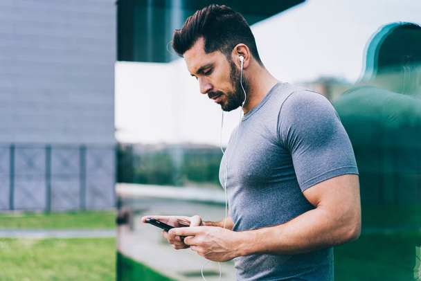 Muscular bodybuilder reading sportive notifications during outdoors workout using 4g internet for listening online music, Caucasian masculine man watching video training for practice in life - Photo, Image
