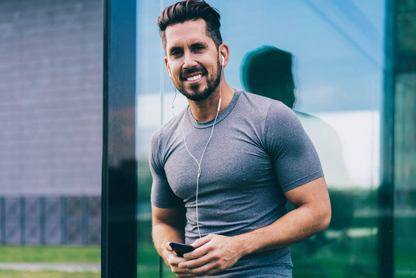 Half length portrait of muscular trainer 30s posing outdoors enjoying music podcast, happy Caucasian bodybuilder in electronic headphones holding modern cellular device and smiling at camera - Foto, afbeelding