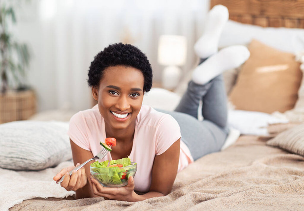 Concepto equilibrado de dieta de alimentos crudos. Hermosa mujer negra comiendo ensalada de verduras frescas en la cama en casa, espacio libre - Foto, Imagen