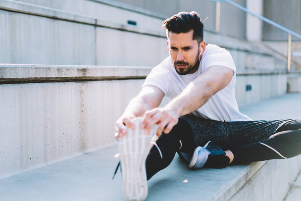 Caucasian muscular guy in tracksuit and earphones have training outdoors keeping wellbeing healthy lifestyle, handsome sportsman doing exercises for legs strength before jogging on morning workout - Foto, Imagem