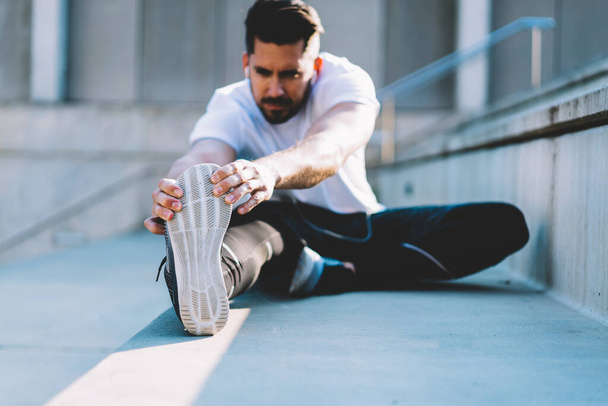 Blurred Caucasian sportsman doing stretching exercise during morning workout at urban setting, selective focus on strength male hands holding sneakers sole doing flexibility warm up with effort - Фото, зображення
