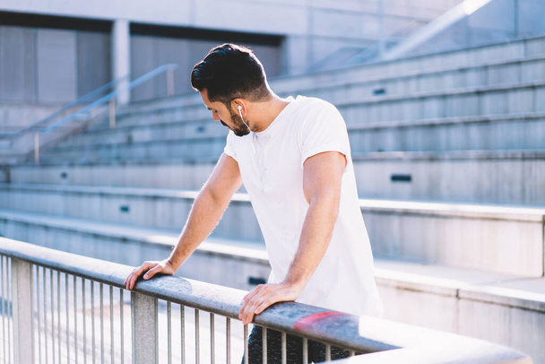 Tired Caucasian male in electronic headphones listening sportive audio podcast during workout break at urban setting, muscular man 30s with perfect body shape resting after training workout - 写真・画像