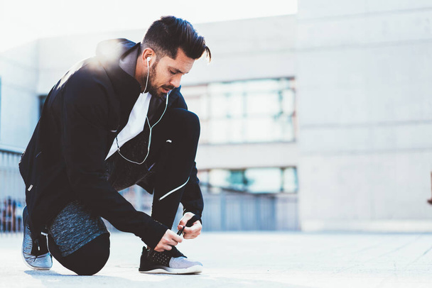 Caucasian sportsman in tracksuit tying laces on sneakers for jogging outdoors, male athlete preparing for cardio workout on street checking cross shoes using earphones for listening motivation music - Фото, зображення
