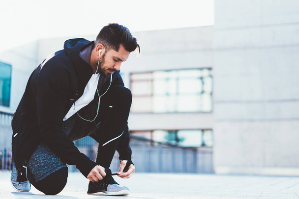 Active sportsman in tracksuit tying laces on sneakers for jogging outdoors, male athlete preparing for cardio workout on street checking cross shoes using earphones for listening motivation music - Foto, afbeelding