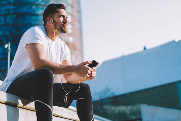 Pensive male bodybuilder in sportswear sitting at urbanity pondering on morning workout exercises, Caucasian athlete with cellphone gadget in hands listening audio podcast during training break - Photo, Image