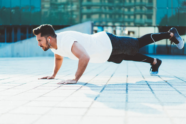 Strong Caucasian sportsman with muscular body shape doing push ups on street enjoying morning workout, strength male athlete in electronic headphones keeping vitality and wellness on activity - Foto, Bild