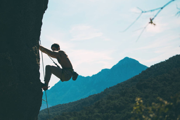 Kalliokiipeilijä kiipeää kalliolle vuorten ja taivaan taustalla, mies kouluttaa voimaa ja kestävyyttä, voittaa korkeiden paikkojen pelon, harjoittelee luonnossa.. - Valokuva, kuva