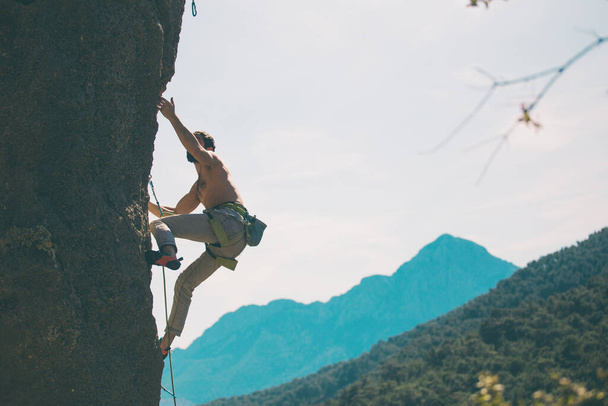 Ein Bergsteiger erklimmt einen Felsen vor dem Hintergrund von Bergen und Himmel, ein Mann trainiert Kraft und Ausdauer, überwindet Höhenangst, trainiert in der Natur. - Foto, Bild