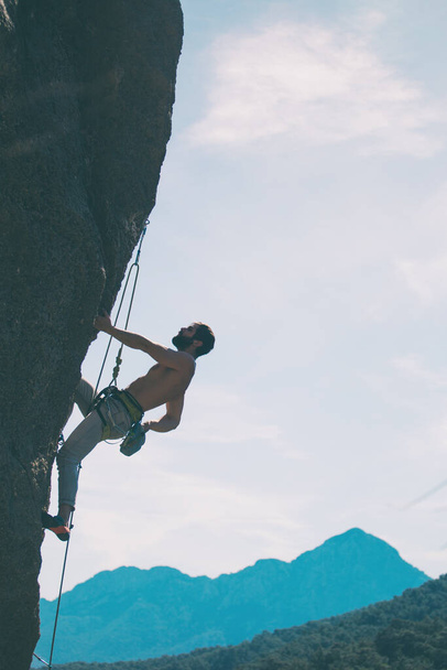 Kalliokiipeilijä kiipeää kalliolle vuorten ja taivaan taustalla, mies kouluttaa voimaa ja kestävyyttä, voittaa korkeiden paikkojen pelon, harjoittelee luonnossa.. - Valokuva, kuva