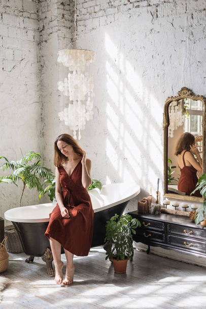 Concept of morning routine. Vertical view smiling young woman sitting on classic bath at comfortable bathroom with green houseplants in flower pots, decor at commode and interior design in boho style - Фото, изображение