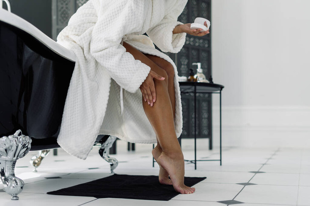 Concept of beauty procedure. Cropped view of afro american woman in bathrobe spending morning in bathroom, applying cream on shaved and soft legs - 写真・画像