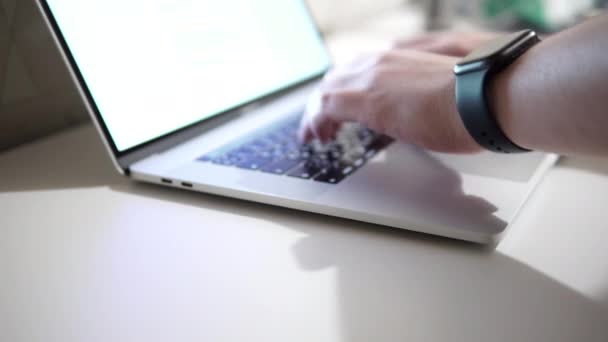 Un hombre con un reloj inteligente en su mano imprime texto, ingresa datos, trabaja con el teclado de un portátil. - Metraje, vídeo