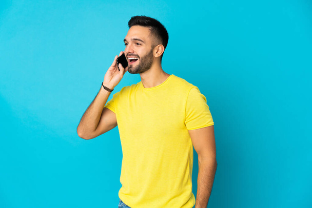 Fiatal fehér férfi elszigetelt kék háttér miközben egy beszélgetést a mobiltelefon - Fotó, kép