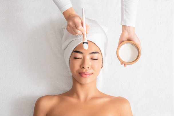 Schönheitsbehandlungen. Kosmetikerin trägt im Wellness-Salon Tonmaske auf asiatisches Frauengesicht auf - Foto, Bild