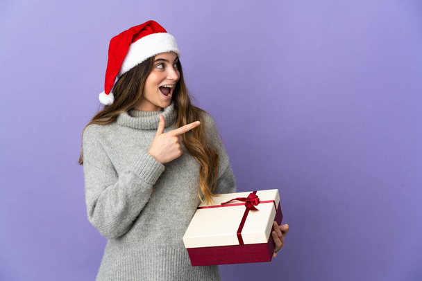 Дівчина з різдвяним капелюхом тримає подарунок ізольовано на білому тлі вказуючи пальцем на бік і представляючи продукт
 - Фото, зображення